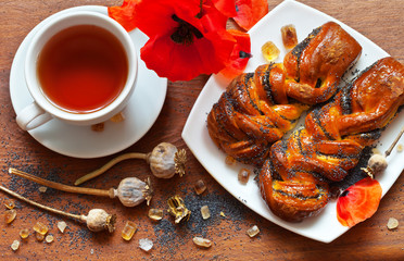 Sweet buns with poppy seeds, red poppy flowers, seed capsules and a cup of tea on a wooden table, top view