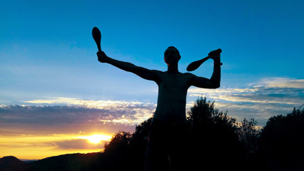 Silhouette of a lean sporty young male exercising outdoor with Indian clubs, with beautiful sunset in the background