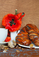 Wicker buns with poppy seeds, porcelain vase with red poppy flowers and a cup of tea on a wooden background closeup