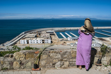 Fototapeta na wymiar Nord Africa: una donna musulmana di spalle, di fronte al porto di Tangeri, guarda lo stretto di Gibilterra che unisce Oceano Atlantico e Mar Mediterraneo e separa il Marocco dalla Spagna