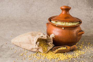   A small linen sack with raw millet and a clay pot with hot porridge on a gray background. Traditional Russian cuisine. The concept of a healthy diet.
