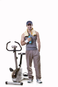 Portrait of a young man with water bottle standing next to an exercise bike over white background 