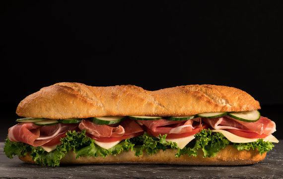 Huge crispy baguette deli sandwich with meat and vegetables. Close up. Black background.