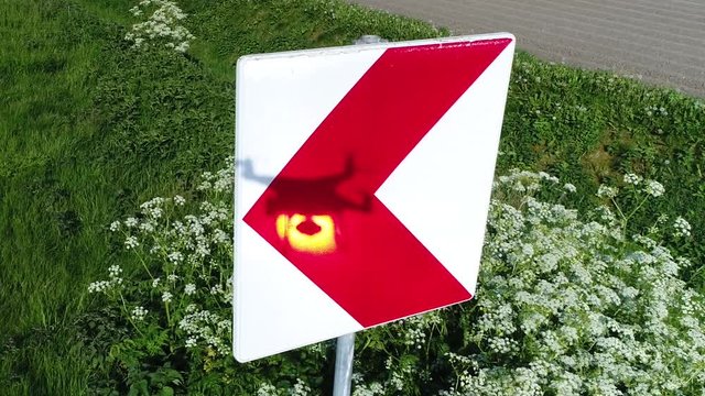 Aerial of road sign red arrow left signal showing shadow of hoovering drone and reflection of sun on sign post around the sign is growing grass and weed footage could indicate drone regulations 4k