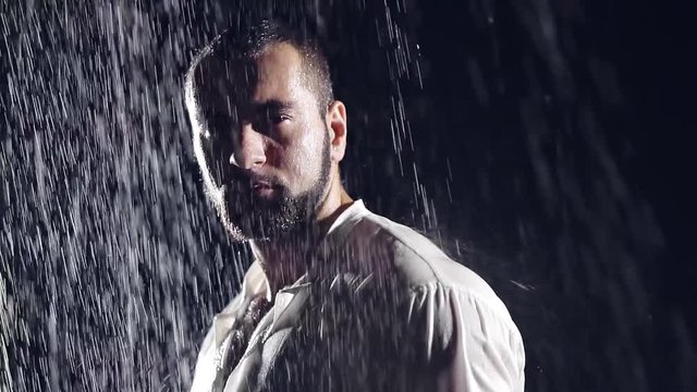 brutal big man with a beard in a white shirt standing on the street at night in the pouring rain soaking wet. sad mood on his face