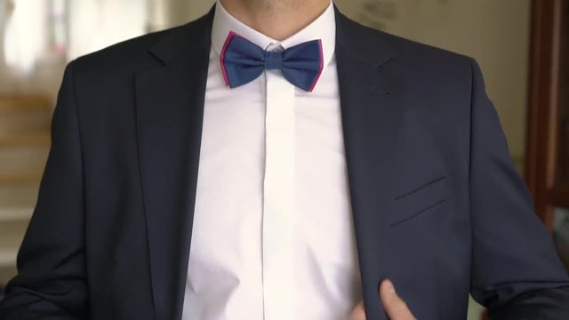 Young man in white shirt with cufflinks fixes purple bow-tie on a neck and buttons and tucks down suit jacket before the wedding ceremony. Interior. Medium.