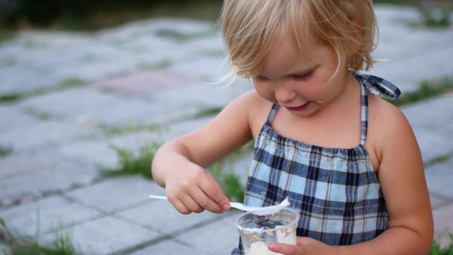 The poor girl sits on the sidewalk and eats from a plastic cup.