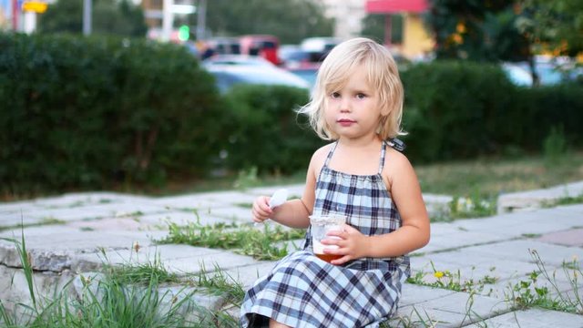 The poor girl sits on the sidewalk and eats from a plastic cup.