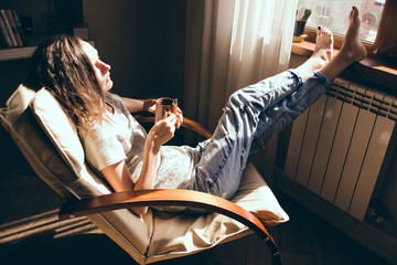 Weekends finally. Woman in blue jeans relaxing with cup of tea in armchair at home, daydreaming. Girl enjoying life, bright sunlight and heat from radiator. Cozy home. Heating season. Mortgage concept