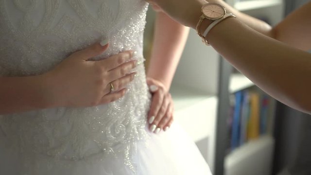 The Bridesmaid Helps Bride to Wear a Wedding Dress, while bride has her right hand with engagement ring on her stomack and lef hand on her waist. Indoor. Sunlight. Move