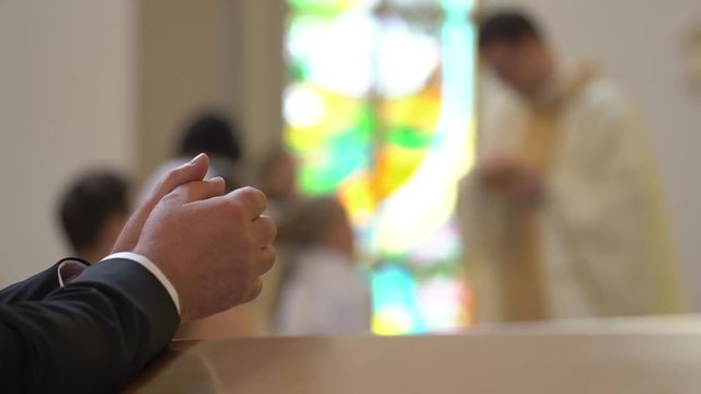 Clasped hands of man kneeling at the pew in the Church and praying. On the blurred background the catholic priest gives the holly communion to people who moving in line. Closed up. Steady. Interior