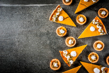 Traditional autumn dishes. Halloween, Thanksgiving. Set of spicy pumpkin pie and pumpkin tartlets with whipped cream & pumpkin seeds on black stone table. top view