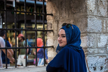 Young and beautiful caucasian covered muslim girl with blue hijab and sunglasses looking through black metal bars near mosque with old stone wall and people walking in the background