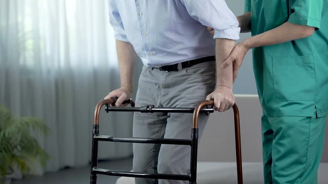 Male patient of nursing home moving with walking frame and nurse, support