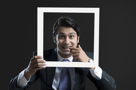 Portrait of young businessman with finger in mouth holding frame against black background