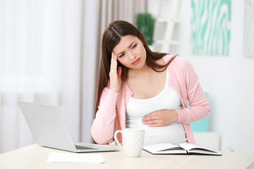 Fototapeta premium Beautiful young pregnant woman suffering from headache while working with laptop at home