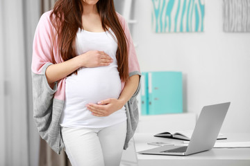 Young pregnant woman standing near table with laptop at home