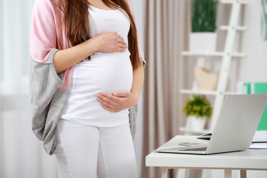Young pregnant woman standing near table with laptop at home
