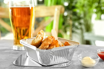 Foil container with tasty fried fish and chips on table
