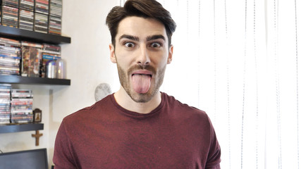 Young Man in Red Shirt Showing Funny Facial Expression with Tongue Out, Indoor at Home