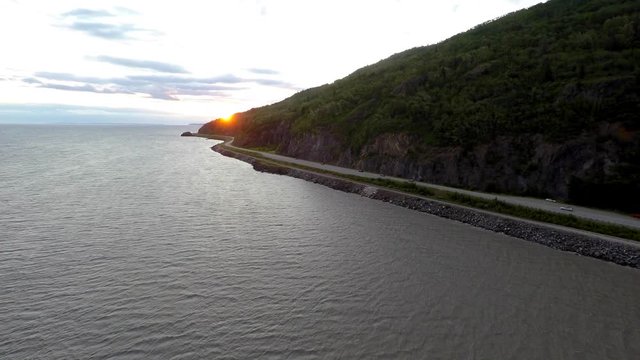 Alaska 4k drone footage of Sunset on Seward Highway at Turnagain Arm tracking over the Cook Inlet with sunset at the point of the mountain side that is touching the water with highway along side.