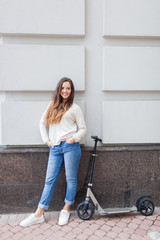 Fototapeta na wymiar Beautiful young girl with long brown hair stopped while riding the scooter on the background of the gray wall. She is dressed in a white sweater and blue jeans