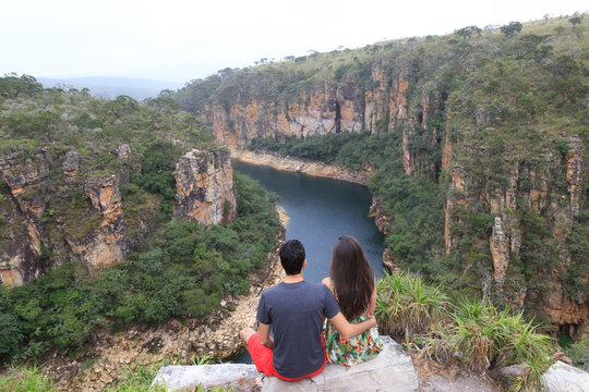 Couple on a rock overlooking a canyon with a river on the bottom and rocky walls covered by green trees. Furnas Canyon is a common tourist destination in Brazil