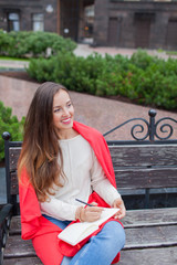 Fototapeta premium An attractive girl with long brown hair and a white tooth smile sits on a bench and writes her thoughts on the urban background in a red notebook. She is wearing a white sweater and blue jeans