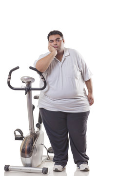 Full length of a tired obese man standing next to an exercise bike over white background 
