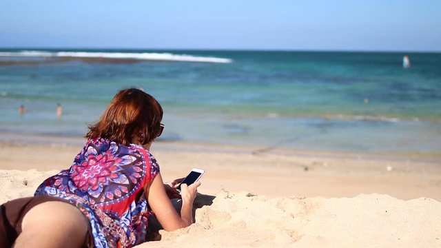 Woman using mobile cell smart phone laughing on beach. Girl in bikini using smartphone happy. Tropical Bali island, Indonesia.