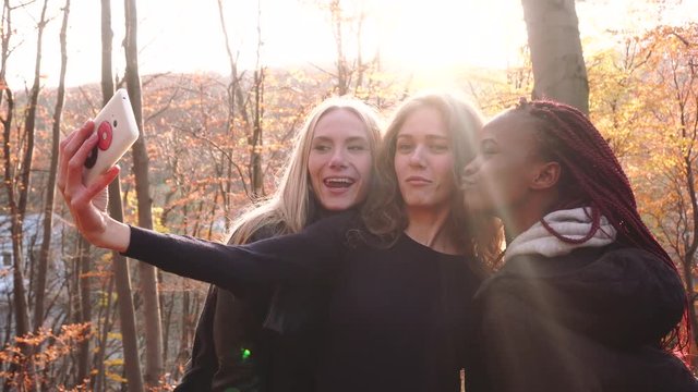 The side view of the multirace girlfriends taking funny selfies in the autumn park.