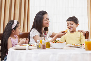 Cheerful mother feeding her son pizza with daughter sitting besides 