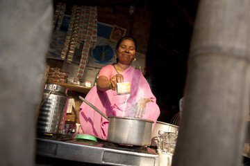 Mature female tea vendor preparing chai on gas stove at market stall 