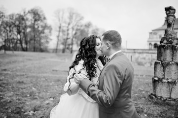 Beautiful wedding couple walking and kissing next to the old fountain in the area with a lovely view of a castle on the background. Rainy weather. Black and white photo.