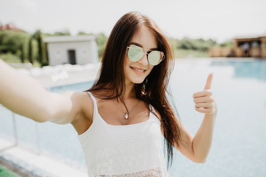 Portrait of beautiful girl taking a selfie at the swimming pool with thumbs up