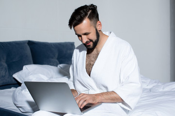 handsome young man in bathrobe using laptop while sitting on bed