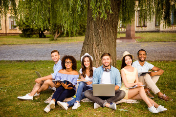 Study together is fun, teamwork,teambuilding concept. Six happy students are sitting under the tree near college building and holding books, devices, wearing casual smart, smiling, on a nice summer