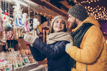 Happy young attractive lady is choosing the gift in a souvenir shop outdoors on a fairy, her lover is embracing her, smiling, they dressed in winter outfits, hats