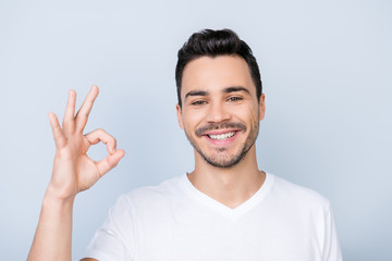 Young cheerful successful guy on the pure light blue background is smiling, wearing casual outfit and is showing ok sign