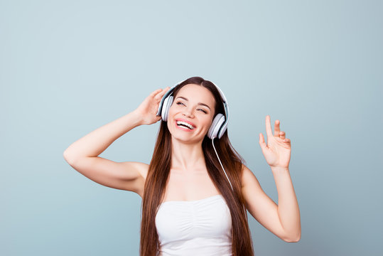 Dreamy charming young lady with toothy smile in modern headphones. She is dreamy and happy, listening to her favourite song, holding the earphones, with closed eyes on pure background