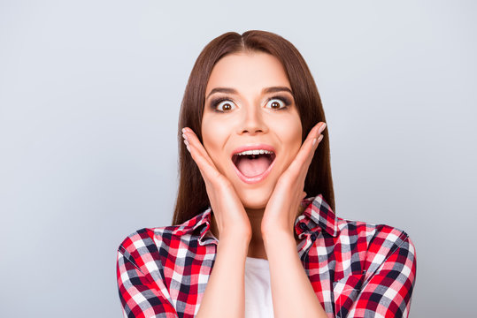 Close up portrait of cute amazed brunette girl in shirt, holding her face, she is shocked, extremely happy, with wide open eyes and mouth on pure background