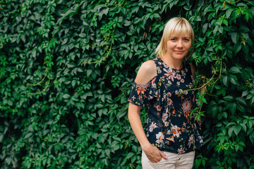 Cute young blonde stylish woman in pretty blouse in flowers over green grape leaves background. Beautiful smiling girl