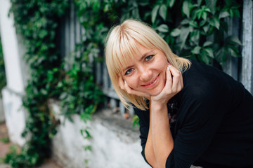 Naklejka premium Cute young blonde beautiful stylish woman in pretty blouse in flowers and black cardigan sitting on fence over green grape leaves background.