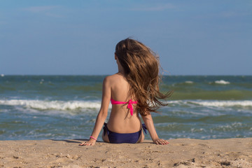 Fototapeta na wymiar Beautiful young woman in a swimsuit with long hair sits on the beach near the sea. Hot summer day