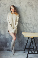 young woman in oversize knitted sweater posing by the grey wall in modern apartment. Casual lifestyle at home