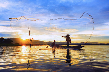 Fisherman on wooden boat casting a net for catching freshwater fish in reservoir at sunrise.