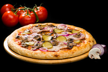 selective focus on appetizing Italian pizza with onion, mushrooms, pickled cucumbers and ham on round wooden board on dark background, near tomatoes and sliced onion