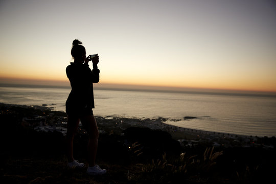 Girl taking photos with phone on beach, silhouette at sunset