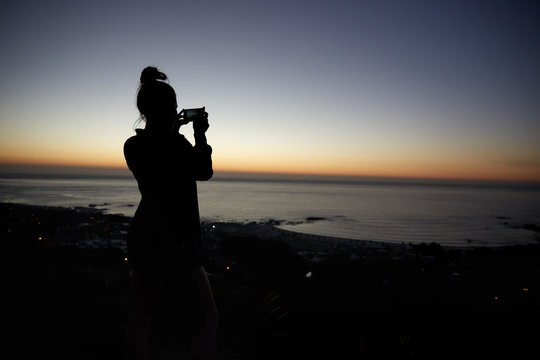 Woman taking photo with phone on beach, silhouette at sunset