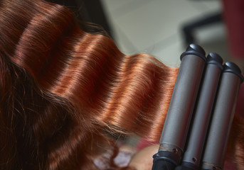 Closeup of long red hair curled with electric triple barrel curling iron in a hairdressing salon.